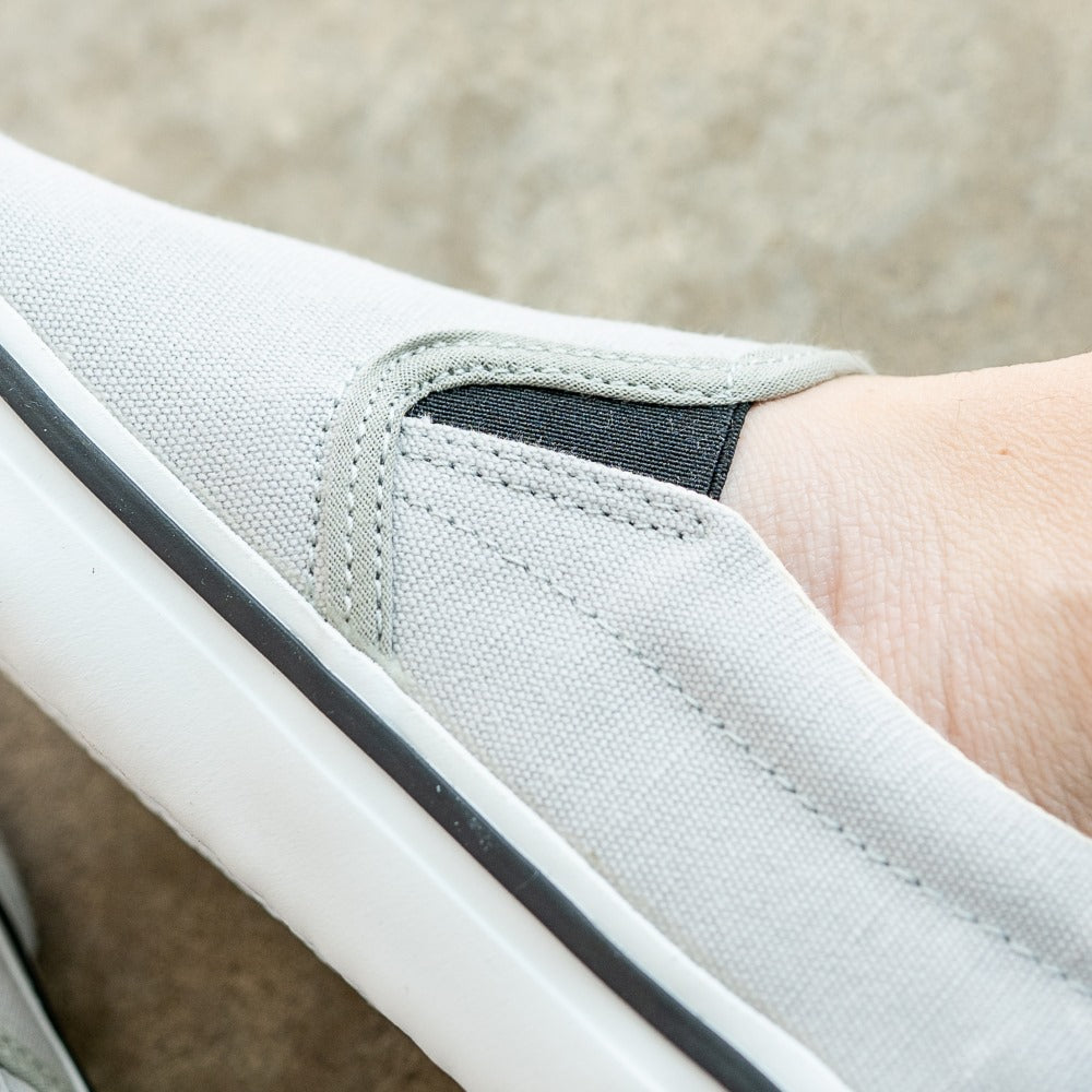 A photo of Bohempia Velik slip on sneakers made from canvas and rubber soles. The sneakers are light grey with white soles with a black stripe around the sole. The right shoe is shown from the left side on a woman's foot, with a close up view of the black elastic detail on the inner top of the foot. #color_light-grey-white