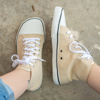 A photo of Bohempia Orik canvas high tops made from canvas and rubber soles. The sneakers are a tan color with a white toe cap and a black outline around the rubber. The left shoe is shown from above and the right shoe is shown from the left side on a woman's feet, with a view of her shins down, on a cement floor. #color_tan-white