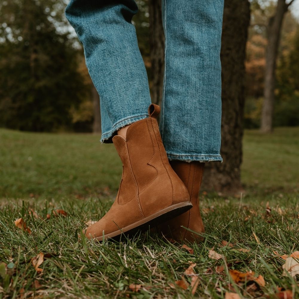 Light brown, nubuck leather western Faye boots By Anya with a western shape on both sides of the ankles, a zipper, tan rubber soles, a welt stitching design around the top of the sole, and a pull tab at the top back of the opening. Both boots are shown facing diagonally to the back left on a light-skinned woman wearing loose, cropped, medium-wash jeans standing in a tree-filled grassy area. #color_cider