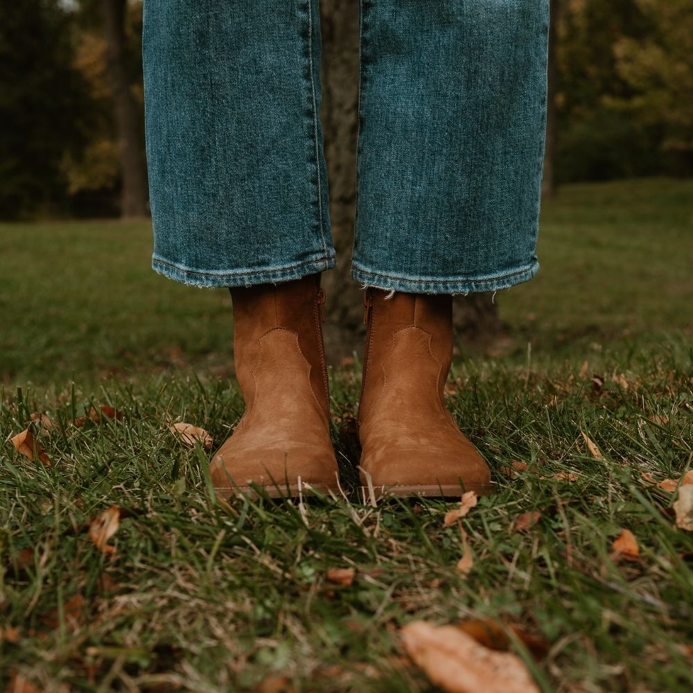 Light brown, nubuck leather western Faye boots By Anya with a western shape on both sides of the ankles, a zipper, tan rubber soles, a welt stitching design around the top of the sole, and a pull tab at the top back of the opening. Both boots are shown from the front on a light-skinned woman wearing loose, cropped, medium-wash jeans standing in a tree-filled grassy area. #color_cider