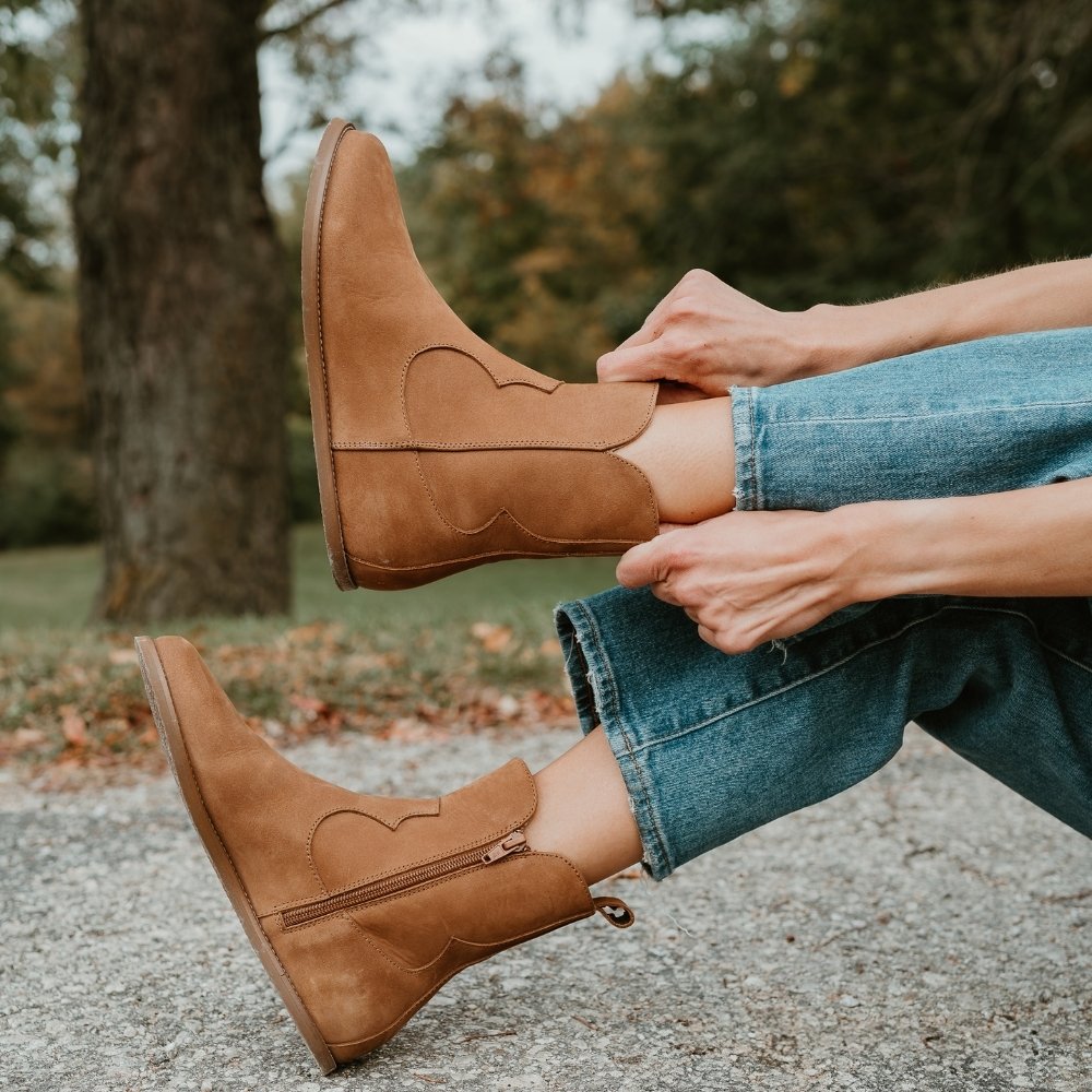 Light brown, nubuck leather western Faye boots By Anya with a western shape on both sides of the ankles, a zipper, tan rubber soles, a welt stitching design around the top of the sole, and a pull tab at the top back of the opening. Both boots are shown from the left pointing up on a light-skinned woman wearing loose, cropped, medium-wash jeans sitting on a paved path holding the left boot. #color_cider