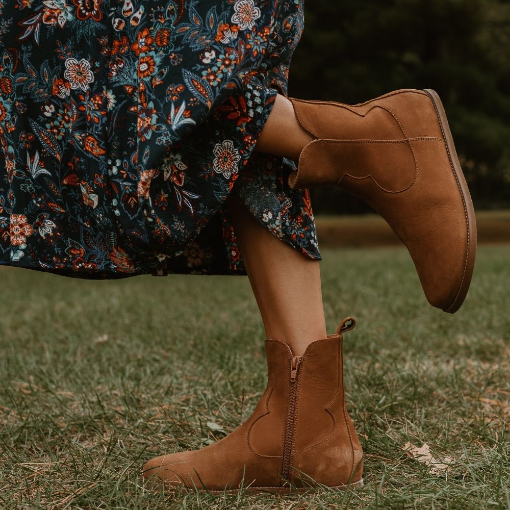 Light brown, nubuck leather western Faye boots By Anya with a western shape on both sides of the ankles, a zipper, tan rubber soles, a welt stitching design around the top of the sole, and a pull tab at the top back of the opening. Both boots are shown facing left on a light-skinned woman wearing a midi, flowy, dark-blue skirt with a white, blue, and red floral, paisley pattern standing in grass. #color_cider