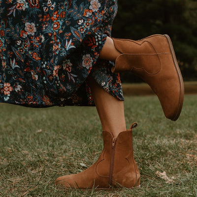 Light brown, nubuck leather western Faye boots By Anya with a western shape on both sides of the ankles, a zipper, tan rubber soles, a welt stitching design around the top of the sole, and a pull tab at the top back of the opening. Both boots are shown facing left on a light-skinned woman wearing a midi, flowy, dark-blue skirt with a white, blue, and red floral, paisley pattern standing in grass. #color_cider
