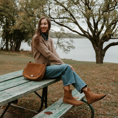 Light brown, nubuck leather western Faye boots By Anya with a western shape on both sides of the ankles, a zipper, tan rubber soles, a welt stitching design around the top of the sole, and a pull tab at the top back of the opening. Both boots are shown facing right on a light-skinned woman with long, brown hair wearing a camel knit sweater, a dark green silk scarf, and loose, cropped, medium-wash jeans sitting on a bench in nature with a brown leather purse. #color_cider
