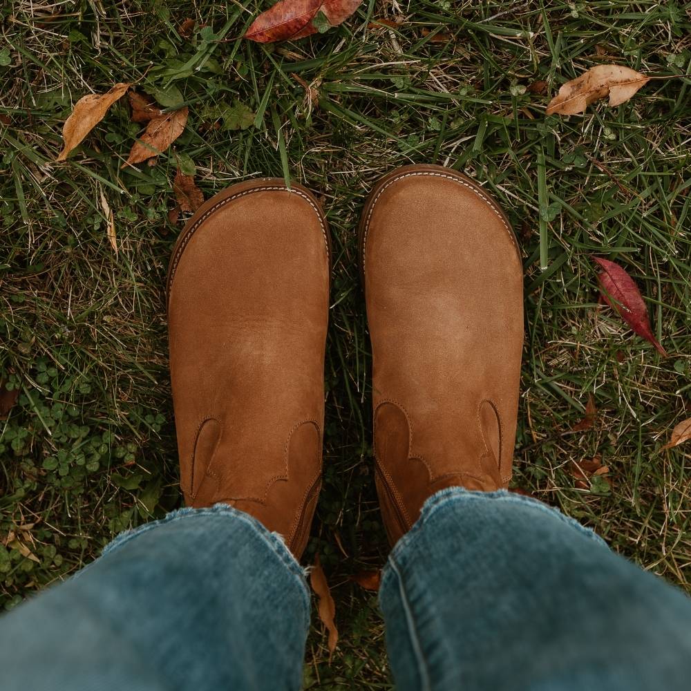Light brown, nubuck leather western Faye boots By Anya with a western shape on both sides of the ankles, a zipper, tan rubber soles, a welt stitching design around the top of the sole, and a pull tab at the top back of the opening. Both boots are shown from above on a woman wearing loose, cropped, medium-wash jeans standing on leafy grass. #color_cider