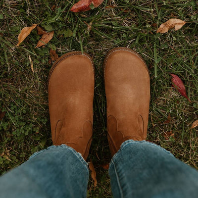 Light brown, nubuck leather western Faye boots By Anya with a western shape on both sides of the ankles, a zipper, tan rubber soles, a welt stitching design around the top of the sole, and a pull tab at the top back of the opening. Both boots are shown from above on a woman wearing loose, cropped, medium-wash jeans standing on leafy grass. #color_cider