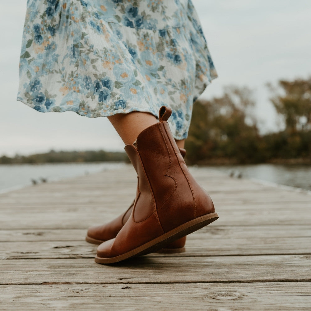 Brown smooth leather western Faye boots By Anya with a western shape on both sides of the ankles, a zipper, tan rubber soles, a welt stitching design around the top of the sole, and a pull tab at the top back of the opening. Both boots are shown facing diagonally to the back left on a light-skinned woman wearing a long, flowy, white dress with a blue & green floral pattern and a knit, ivory button up sweater standing on a dock. #color_cognac