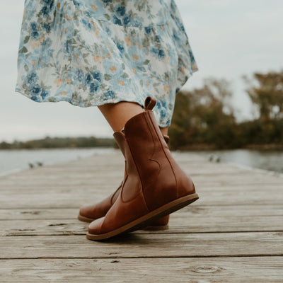 Brown smooth leather western Faye boots By Anya with a western shape on both sides of the ankles, a zipper, tan rubber soles, a welt stitching design around the top of the sole, and a pull tab at the top back of the opening. Both boots are shown facing diagonally to the back left on a light-skinned woman wearing a long, flowy, white dress with a blue & green floral pattern and a knit, ivory button up sweater standing on a dock. #color_cognac