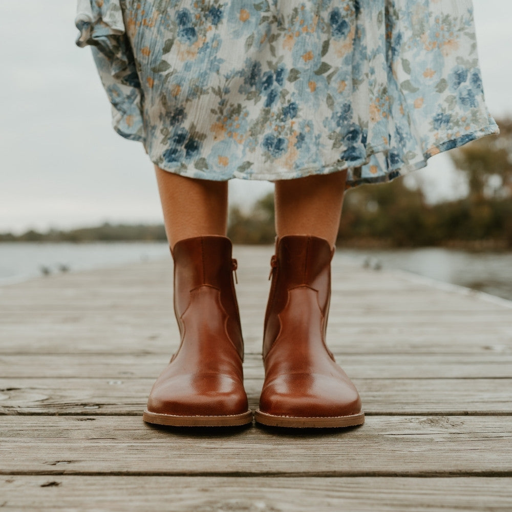 Brown smooth leather western Faye boots By Anya with a western shape on both sides of the ankles, a zipper, tan rubber soles, a welt stitching design around the top of the sole, and a pull tab at the top back of the opening. Both boots are shown from the front on a woman wearing a long, flowy, white dress with a blue & green floral pattern and a knit, ivory button up sweater standing on a dock. #color_cognac