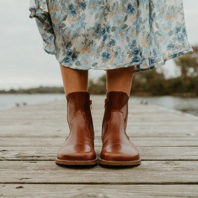 Brown smooth leather western Faye boots By Anya with a western shape on both sides of the ankles, a zipper, tan rubber soles, a welt stitching design around the top of the sole, and a pull tab at the top back of the opening. Both boots are shown from the front on a woman wearing a long, flowy, white dress with a blue & green floral pattern and a knit, ivory button up sweater standing on a dock. #color_cognac
