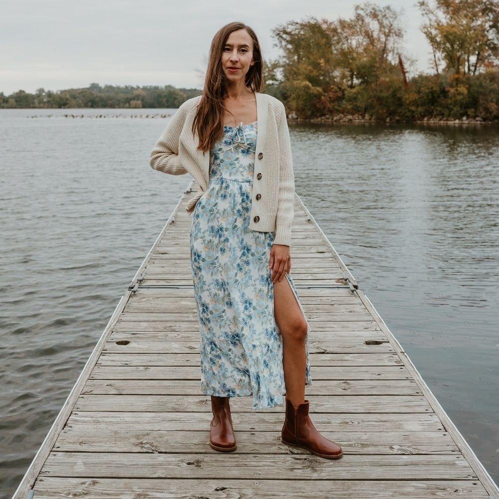 Brown smooth leather western Faye boots By Anya with a western shape on both sides of the ankles, a zipper, tan rubber soles, a welt stitching design around the top of the sole, and a pull tab at the top back of the opening. Both boots are shown from the front on a light-skinned woman with long, brown hair wearing a long, flowy, white dress with a blue & green floral pattern and a knit, ivory button up sweater standing on a dock. #color_cognac