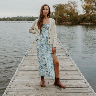 Brown smooth leather western Faye boots By Anya with a western shape on both sides of the ankles, a zipper, tan rubber soles, a welt stitching design around the top of the sole, and a pull tab at the top back of the opening. Both boots are shown from the front on a light-skinned woman with long, brown hair wearing a long, flowy, white dress with a blue & green floral pattern and a knit, ivory button up sweater standing on a dock. #color_cognac