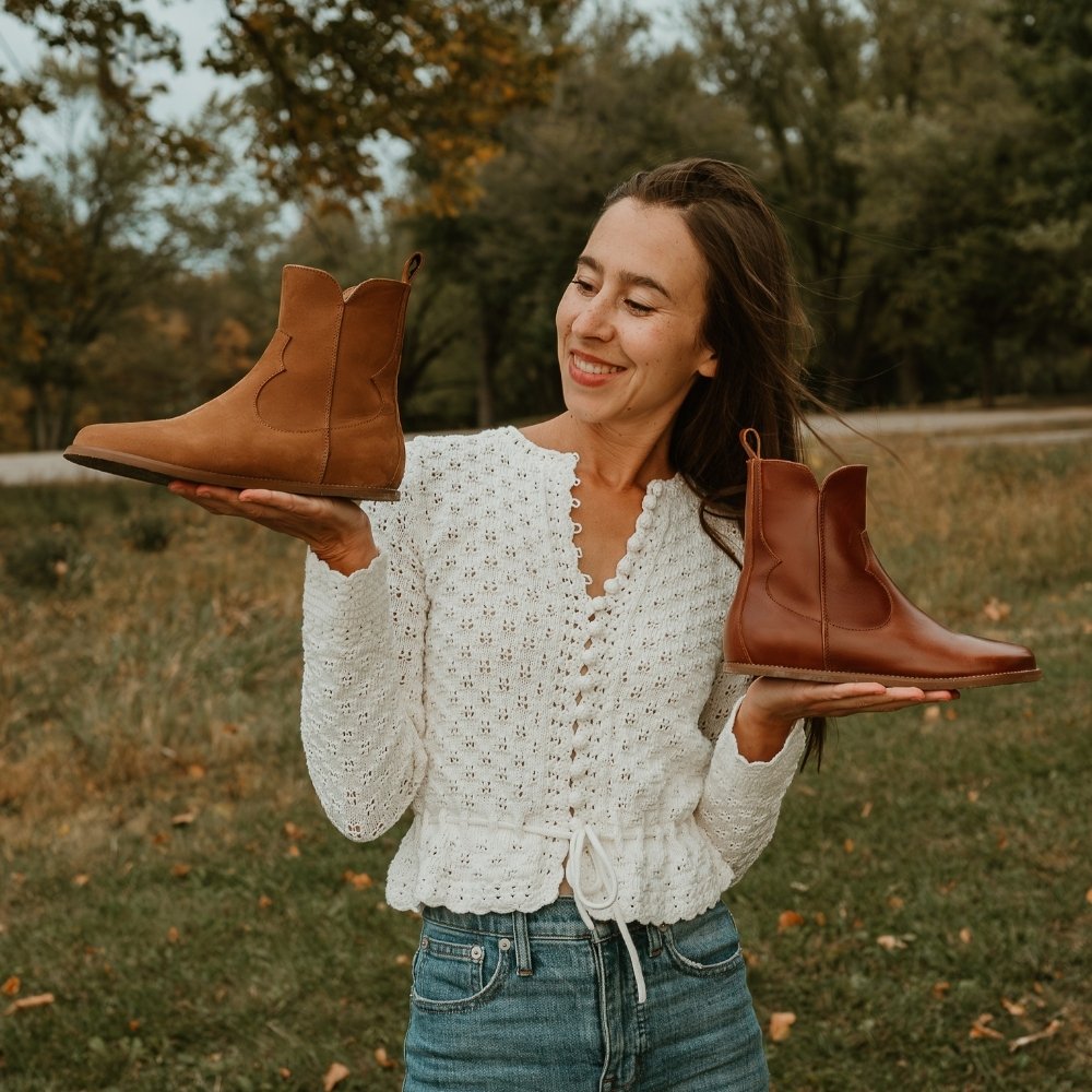 Brown smooth leather western Faye boots By Anya with a western shape on both sides of the ankles, a zipper, tan rubber soles, a welt stitching design around the top of the sole, and a pull tab at the top back of the opening. Cider boot is shown facing left in one hand and cognac boot is shown facing right in the other hand being held by a light-skinned woman with long, brown hair wearing a white, long-sleeve, knitted top, and loose, cropped, medium-wash jeans standing in nature. #color_cider