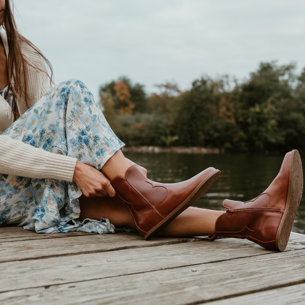 Brown smooth leather western Faye boots By Anya with a western shape on both sides of the ankles, a zipper, tan rubber soles, a welt stitching design around the top of the sole, and a pull tab at the top back of the opening. Both boots are shown from the right pointing upwards on a light-skinned woman with long, brown hair wearing a long, flowy, white dress with a blue & green floral pattern and a knit, ivory button up sweater sitting on a dock holding the right boot. #color_cognac