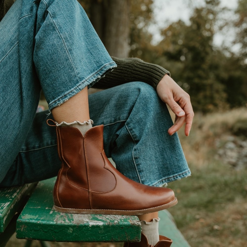 Brown smooth leather western Faye boots By Anya with a western shape on both sides of the ankles, a zipper, tan rubber soles, a welt stitching design around the top of the sole, and a pull tab at the top back of the opening. Right boot is shown facing right on a light-skinned woman wearing a dark green knit sweater, loose, cropped, medium-wash jeans sitting on a bench in nature. #color_cognac