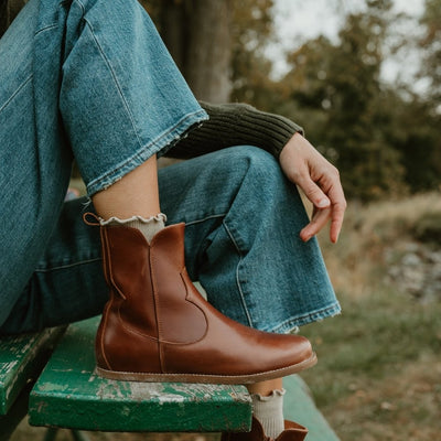 Brown smooth leather western Faye boots By Anya with a western shape on both sides of the ankles, a zipper, tan rubber soles, a welt stitching design around the top of the sole, and a pull tab at the top back of the opening. Right boot is shown facing right on a light-skinned woman wearing a dark green knit sweater, loose, cropped, medium-wash jeans sitting on a bench in nature. #color_cognac