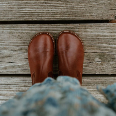 Brown smooth leather western Faye boots By Anya with a western shape on both sides of the ankles, a zipper, tan rubber soles, a welt stitching design around the top of the sole, and a pull tab at the top back of the opening. Both boots are shown from above on a woman wearing a long, flowy, white dress with a blue & green floral pattern and a knit, ivory button up sweater standing on a dock. #color_cognac