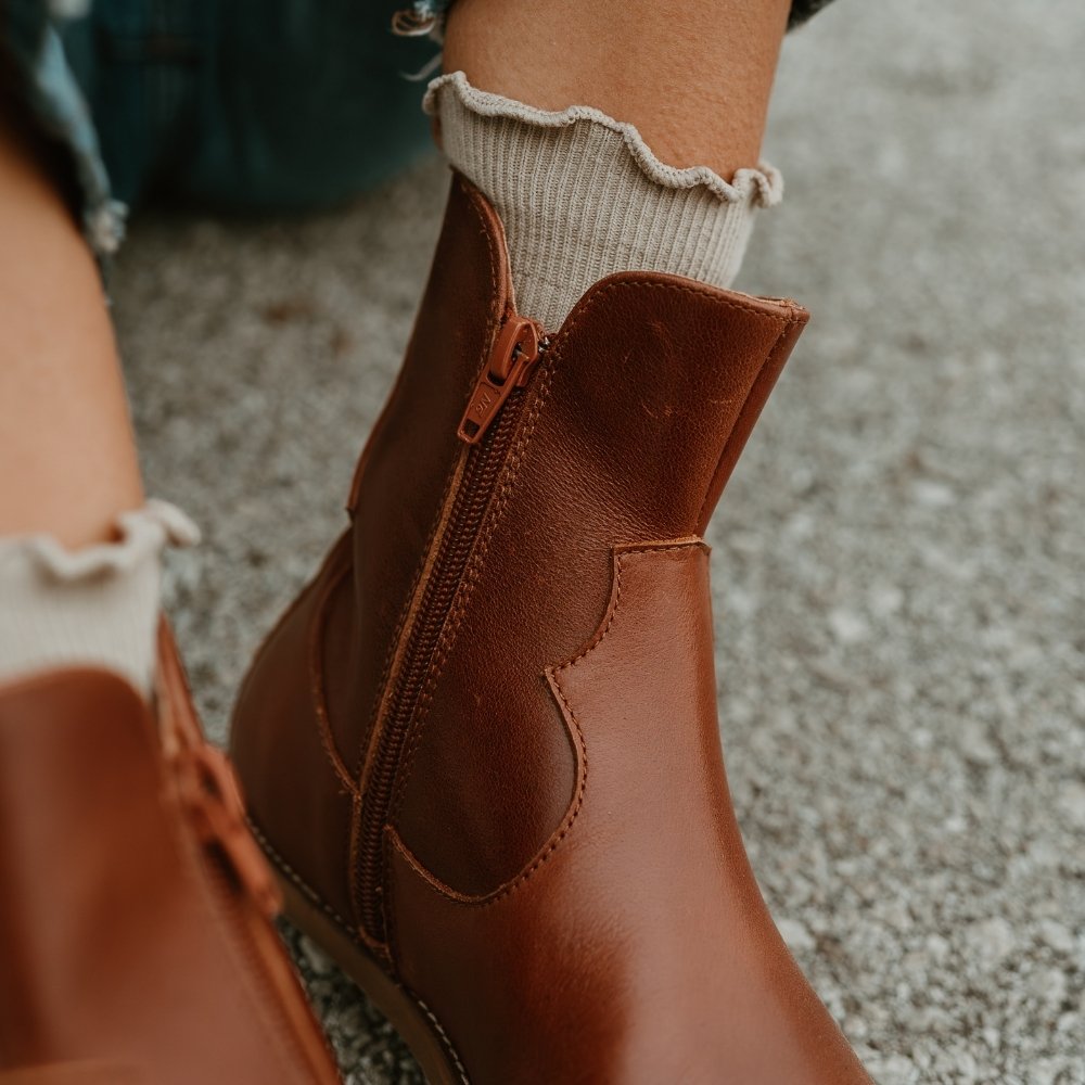 Brown smooth leather western Faye boots By Anya with a western shape on both sides of the ankles, a zipper, tan rubber soles, a welt stitching design around the top of the sole, and a pull tab at the top back of the opening. Right boot is shown close up to the zipper on a light-skinned woman wearing loose, cropped, medium-wash jeans and tan socks sitting on pavement. #color_cognac