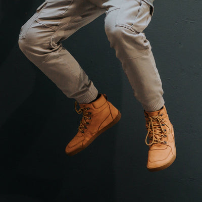 Cognac Crave Glasgow leather boots. Boots are a work-type boot with durable round yellow laces and two quick lace loops at the ankles. Both shoes are shown facing diagonally left on a man jumping wearing grey cargo joggers against a charcoal background. #color_cognac