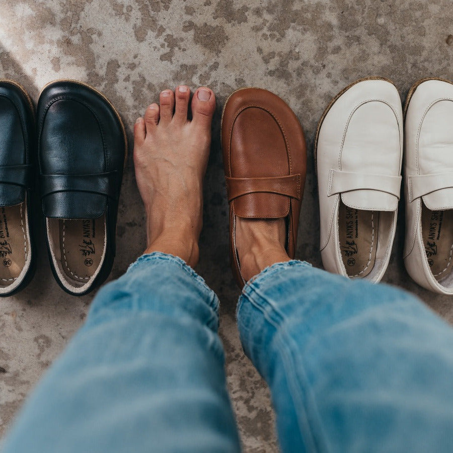 A photo of Dalia Leather loafers Designed by Anya with a leather upper and tan rubber soles. The loafers are a smooth leather upper and have a matching leather strap across the top of the foot for design. Three pairs of loafers are shown from above: a black pair on the left, brown in the middle, and beige on the right. A woman is wearing the right brown loafer with her bare left foot next to it. She is wearing cropped blue jeans and the view is of her knees down. #color_black