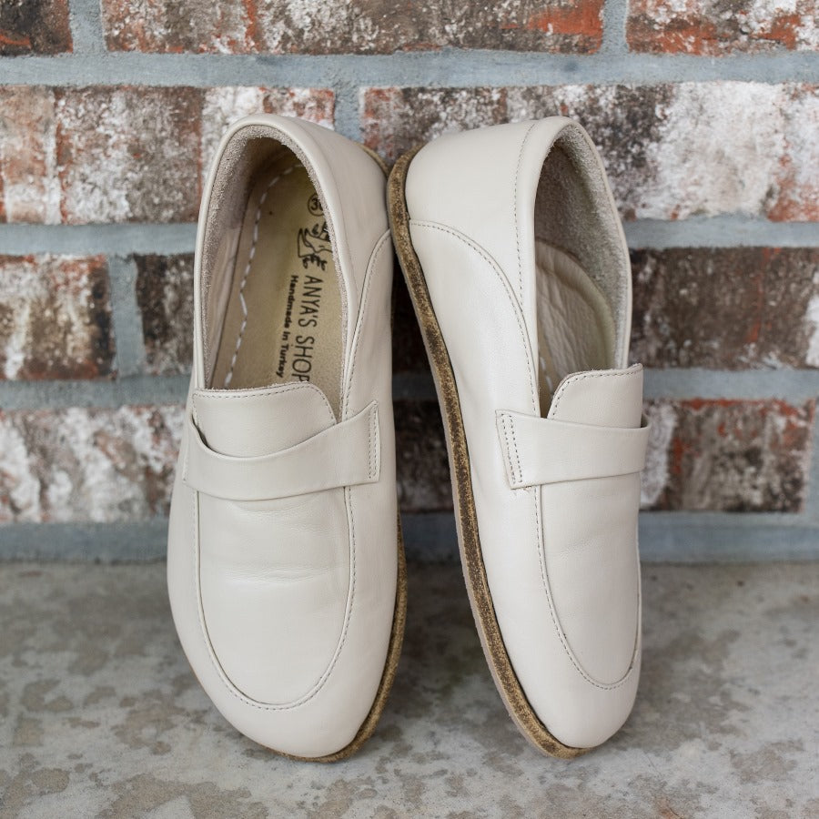 A photo of Dalia Leather loafers Designed by Anya with a leather upper and tan rubber soles. The loafers are a beige smooth leather upper and have a matching leather strap across the top of the foot for design. Both loafers are shown beside each other propped against a brick wall, with the front of the right shoe and the right side of the left shoe shown. #color_beige