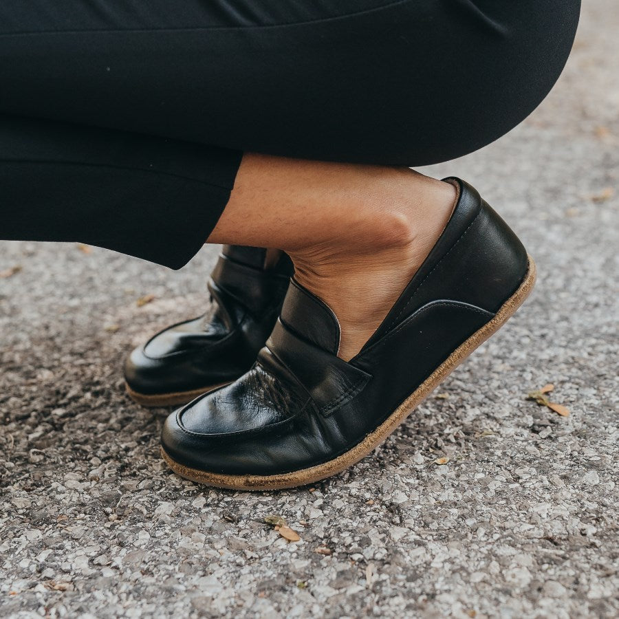 A photo of Dalia Leather loafers Designed by Anya with a leather upper and tan rubber soles. The loafers are a black smooth leather upper and have a matching leather strap across the top of the foot for design. Both loafers are shown from the left side on a woman’s feet, with a view of her waist down. The woman is wearing skinny black pants and the loafers, and is sitting on her heels to demonstrate the flexibility of the soles. #color_black
