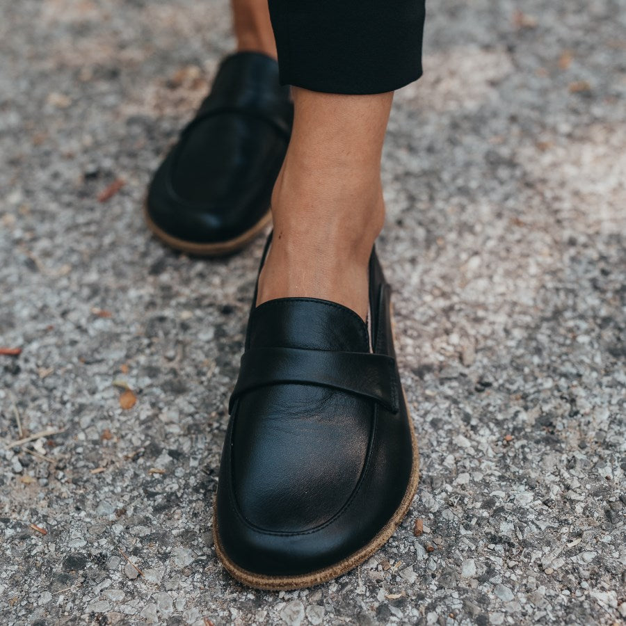 A photo of Dalia Leather loafers Designed by Anya with a leather upper and tan rubber soles. The loafers are a black smooth leather upper and have a matching leather strap across the top of the foot for design. Both loafers are shown from the front on a woman’s feet, with a view of her ankles down. The woman is wearing skinny black pants and the loafers, and is standing on asphalt with her left foot in front of her right. #color_black