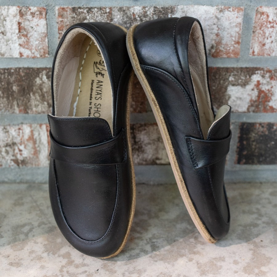 A photo of Dalia Leather loafers Designed by Anya with a leather upper and tan rubber soles. The loafers are a black smooth leather upper and have a matching leather strap across the top of the foot for design. Both loafers are shown beside each other propped against a brick wall, with the front of the right shoe and the right side of the left shoe shown. #color_black