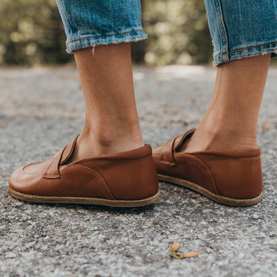 A photo of Dalia Leather loafers Designed by Anya with a leather upper and tan rubber soles. The loafers are a brown smooth leather upper and have a matching leather strap across the top of the foot for design. Both loafers are shown from the left side on a woman’s feet with a view of her shins down. The woman is wearing cropped blue jeans and is standing on an asphalt road with her left foot slightly in front of her right. #color_brown