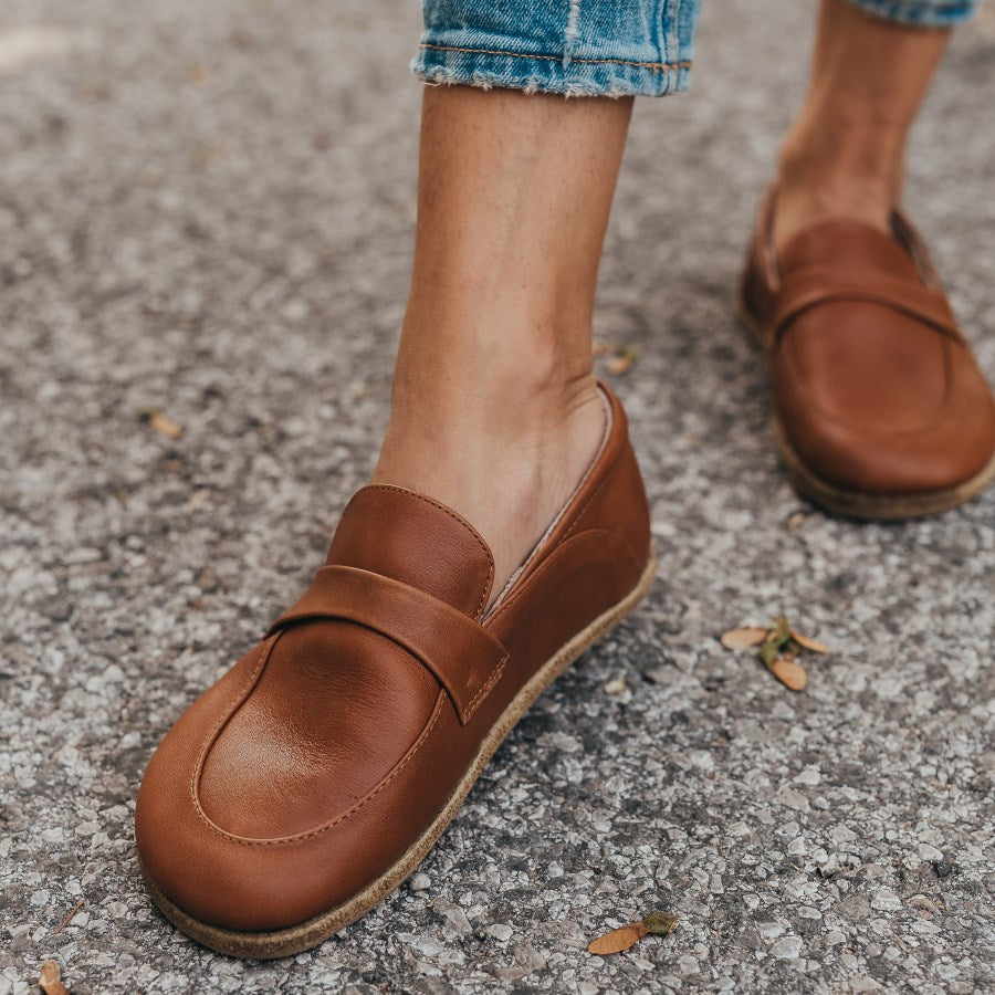 A photo of Dalia Leather loafers Designed by Anya with a leather upper and tan rubber soles. The loafers are a brown smooth leather upper and have a matching leather strap across the top of the foot for design. Both loafers are shown from the front on a woman’s feet with a view of her shins down. The woman is wearing cropped blue jeans and is standing on an asphalt road with her right foot in front of her left. #color_brown