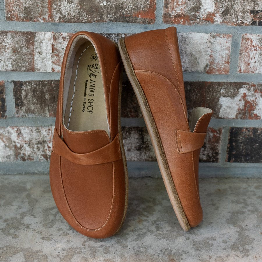 A photo of Dalia Leather loafers Designed by Anya with a leather upper and tan rubber soles. The loafers are a brown smooth leather upper and have a matching leather strap across the top of the foot for design. Both loafers are shown beside each other propped against a brick wall, with the front of the right shoe and the right side of the left shoe shown. #color_brown