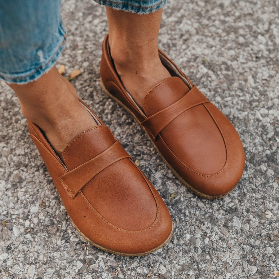 A photo of Dalia Leather loafers Designed by Anya with a leather upper and tan rubber soles. The loafers are a brown smooth leather upper and have a matching leather strap across the top of the foot for design. Both loafers are shown from above on a woman’s feet with a view of her shins down. The woman is wearing cropped blue jeans and is standing on an asphalt road. #color_brown 