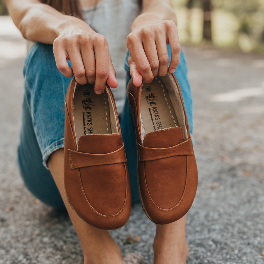 A photo of Dalia Leather loafers Designed by Anya with a leather upper and tan rubber soles. The loafers are a brown smooth leather upper and have a matching leather strap across the top of the foot for design. A woman is sitting on an asphalt road with her knees up, and she is holding both loafers out, displaying what they look like from above. She is wearing cropped blue jeans and a tan shirt. #color_brown