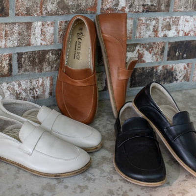 A photo of Dalia Leather loafers Designed by Anya with a leather upper and tan rubber soles. The loafers are a smooth leather upper and have a matching leather strap across the top of the foot for design. Three pairs of loafers are shown: a beige pair on the left shown from the right side, a brown pair in the middle propped against a brick wall, and black pair on the right facing forward. #color_beige