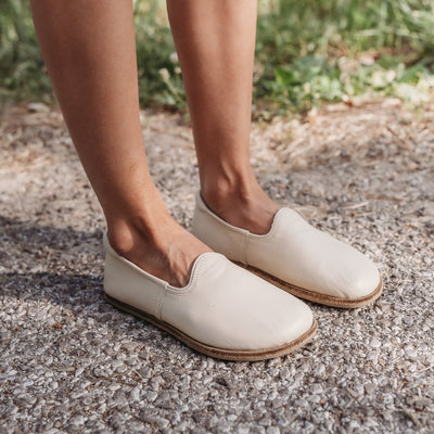 A photo of Yasemin Leather loafers Designed by Anya with a leather upper and tan rubber soles. The loafers are a smooth beige color and have a small curve up on the top of the foot for design. Both loafers shown on a person facing diagonally right standing on pavement. #color_beige