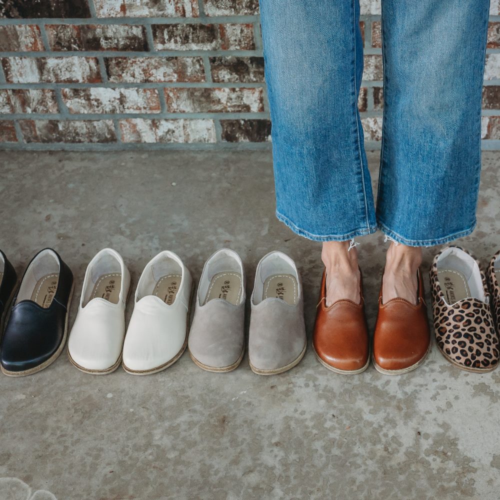 A photo of Yasemin Leather loafers Designed by Anya with a leather upper and tan rubber soles. The loafers have a small curve up on the top of the foot for design. Black, beige, taupe, brown, and leopard loafers shown in a ilne. Brown loafers are on a person wearing loose, cropped, light wash jeans standing on cement with a brick wall in the background. #color_black-smooth-leather