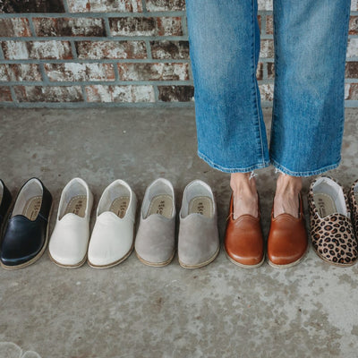 A photo of Yasemin Leather loafers Designed by Anya with a leather upper and tan rubber soles. The loafers have a small curve up on the top of the foot for design. Black, beige, taupe, brown, and leopard loafers shown in a ilne. Brown loafers are on a person wearing loose, cropped, light wash jeans standing on cement with a brick wall in the background. #color_leopard-print-calf-hair