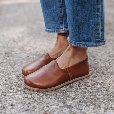 A photo of Yasemin Leather loafers Designed by Anya with a leather upper and tan rubber soles. The loafers are a smooth brown color and have a small curve up on the top of the foot for design. Both loafers shown on a person wearing loose, cropped, light wash jeans facing diagonally left standing on pavement. #color_brown