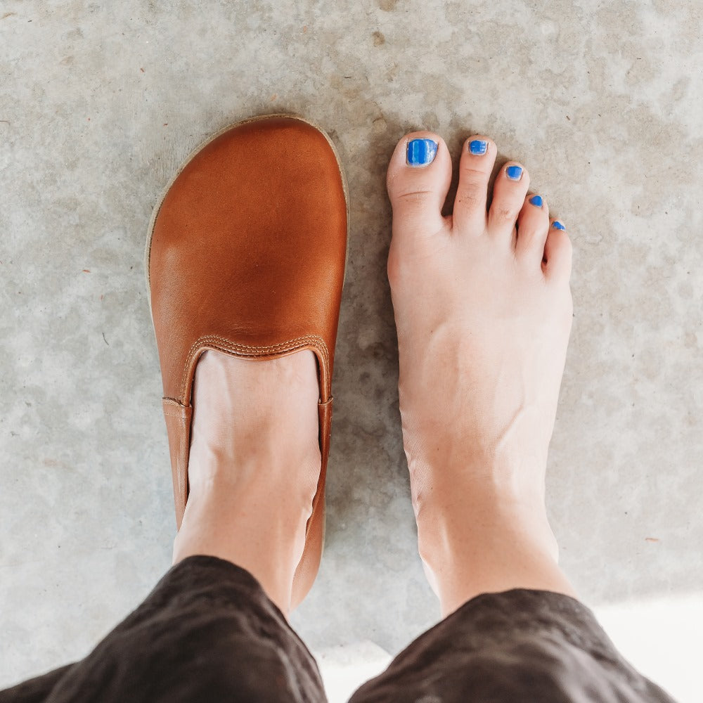 A photo of Yasemin Leather loafers Designed by Anya with a leather upper and tan rubber soles. The loafers are a smooth brown color and have a small curve up on the top of the foot for design. Left loafer is shown from above on a foot next to a bare right foot standing on cement. #color_brown