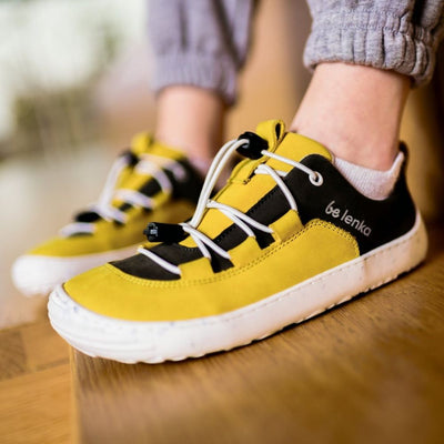 A photo of Be Lenka Xplorer kids shoes in yellow and olive black nubuck leather with white soles and white laces. The black color goes from the heel to the front of the shoe with a yellow tongue, pull tab, and around the side and front edges of the toe box. Elastic laces are laced through yellow tabs attached to the yellow leather accents on sides and end at the black color on either side of the tongue. #color_yellow-olive-black