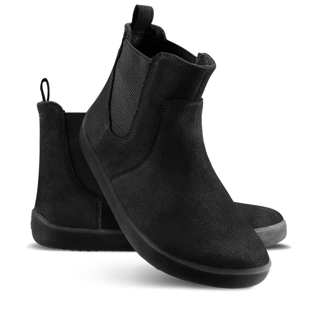 A photo of Be Lenka Entice boots made from nubuck leather and rubber soles. The boots are black in color, they are a Chelsea boot style with elastic at the sides. Left shoe is shown from the right side with the left boot propped up against it with a white background. #color_matte-black