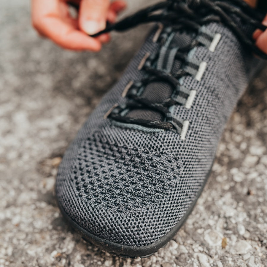 A photo of Freet Pace sneakers in charcoal. Sneakers are completely knit and have a detatched tongue. Laces are black and loop through fabric tabs attached to the shoe and a tab on the tongue. Right shoe is shown close up to the toe box here diagonally from the left side on pavement. #color_charcoal