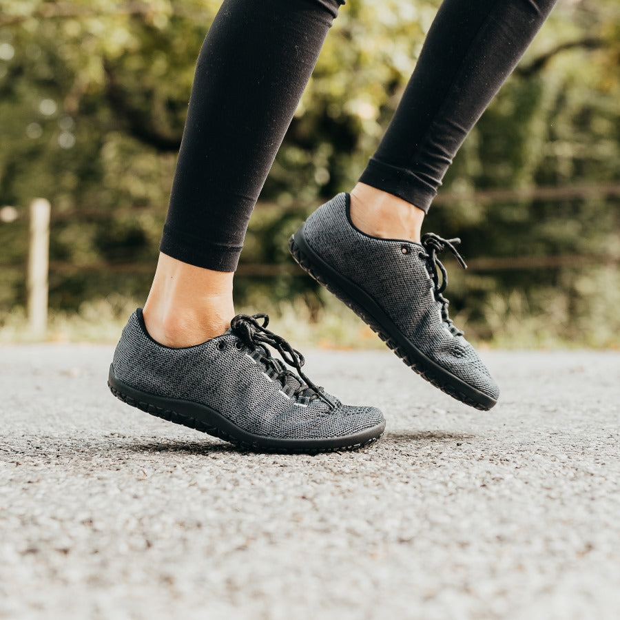 A photo of Freet Pace sneakers in charcoal. Sneakers are completely knit and have a detatched tongue. Laces are black and loop through fabric tabs attached to the shoe and a tab on the tongue. Both shoes are shown here from the right side on a woman's feet running on pavement with woods in the background. #color_charcoal