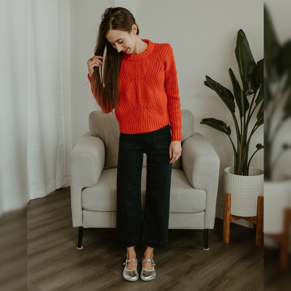 Silver Groundies Mary Jane with silver strap buckle and grey rubber soles. Both shoes are shown facing forward on a light skinned woman wearing a reddish orange sweater and cropped, wide leg, dark wash jeans standing on a grey wooden floor with a light grey chair and plant in the background. #color_silver