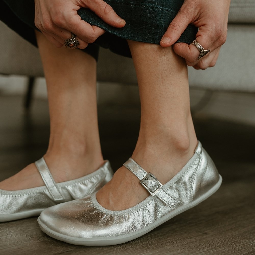 Silver Groundies Mary Jane with silver strap buckle and grey rubber soles. Both shoes are shown facing left on a light skinned woman wearing cropped, wide leg, dark wash jeans sitting on a chair on a grey wooden floor. #color_silver