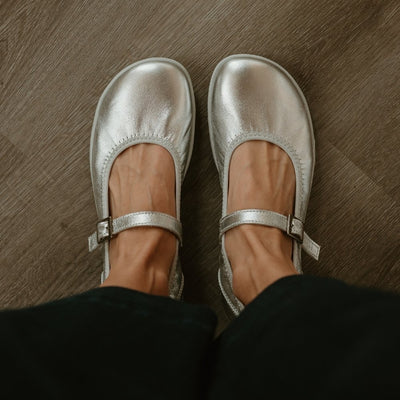 Silver Groundies Mary Jane with silver strap buckle and grey rubber soles. Both shoes are shown from above on a light skinned woman wearing cropped, wide leg, dark wash jeans standing on a grey wooden floor. #color_silver