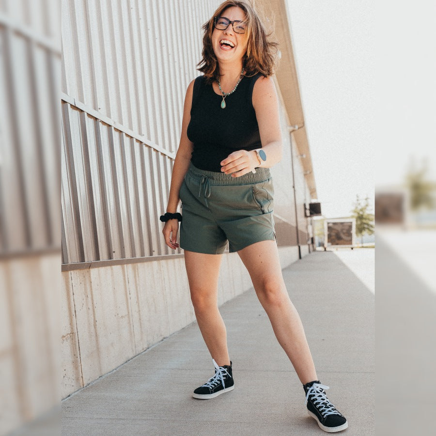 A photo of Groundies Brooklyn Hi-Top sneakers made with canvas and a white rubber sole. The sneakers have a high-top that goes around the ankle with white laces that go from the back end of the toe box all the way up. Both sneakers are shown from the front being worn by a woman with brown hair and glasses. The woman is laughing and standing on a paved sidewalk in front of an industrial looking building, and she is wearing olive green shorts with a black tank top. #color_black