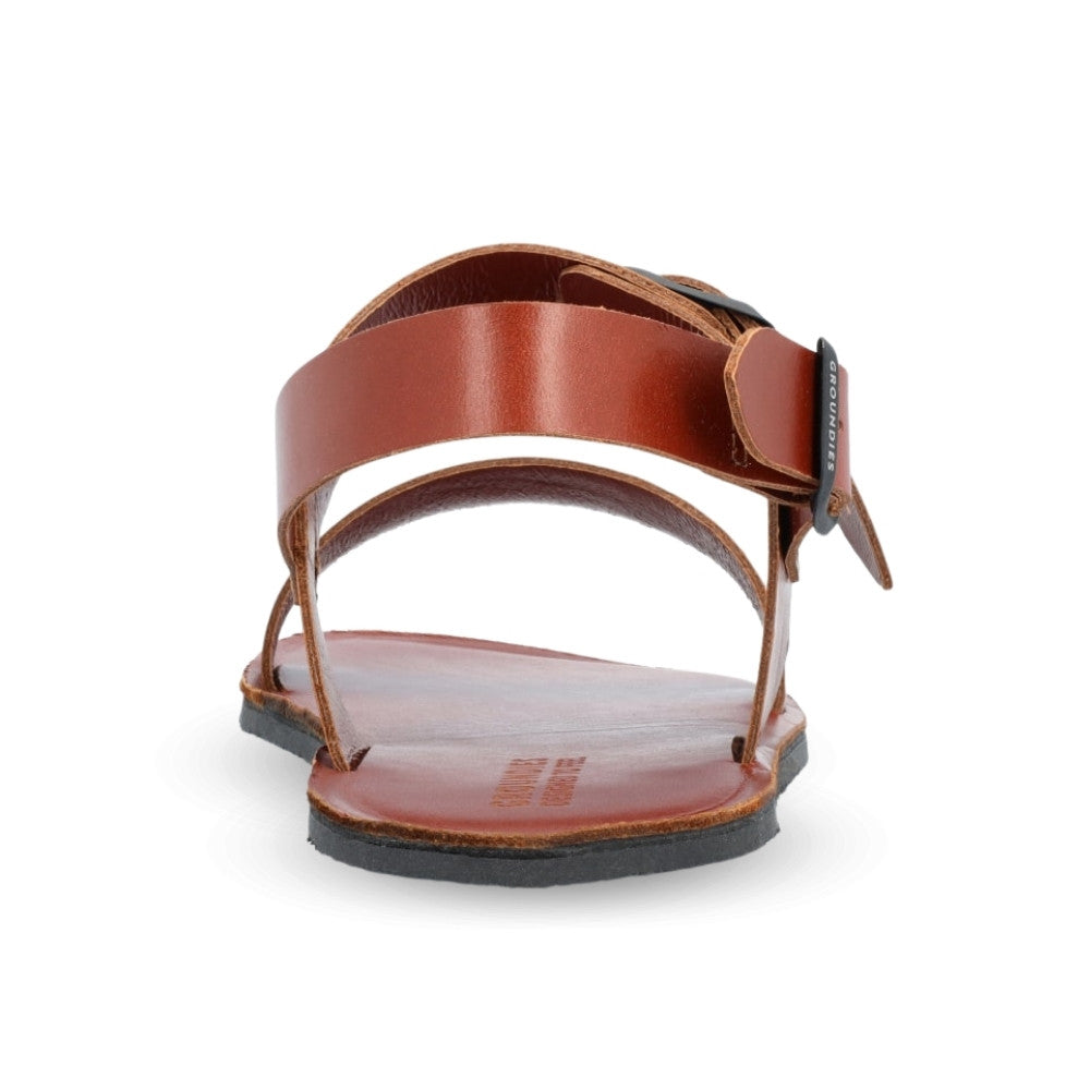 Brown leather Groundies Kos Sandals with black rubber soles. One thick strap spans across the base of the toes while another goes across the arch of the foot with a big, black buckle closure. A thinner slingback strap keeps the sandals secure to the foot also with a buckle closure. Right shoe is shown from behind against a white background. #color_cognac