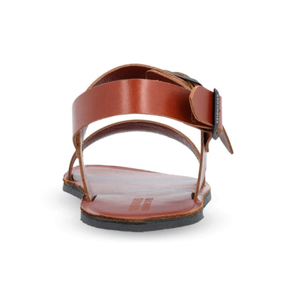 Brown leather Groundies Kos Sandals with black rubber soles. One thick strap spans across the base of the toes while another goes across the arch of the foot with a big, black buckle closure. A thinner slingback strap keeps the sandals secure to the foot also with a buckle closure. Right shoe is shown from behind against a white background. #color_cognac