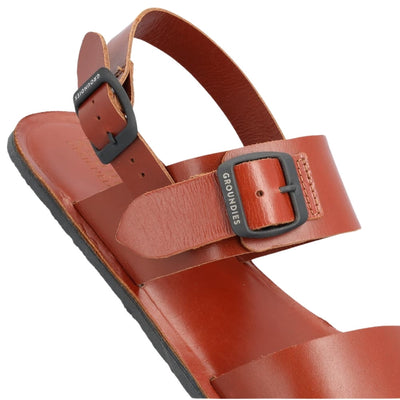 Brown leather Groundies Kos Sandals with black rubber soles. One thick strap spans across the base of the toes while another goes across the arch of the foot with a big, black buckle closure. A thinner slingback strap keeps the sandals secure to the foot also with a buckle closure. Right shoe back buckles are shown close up against a white background. #color_cognac